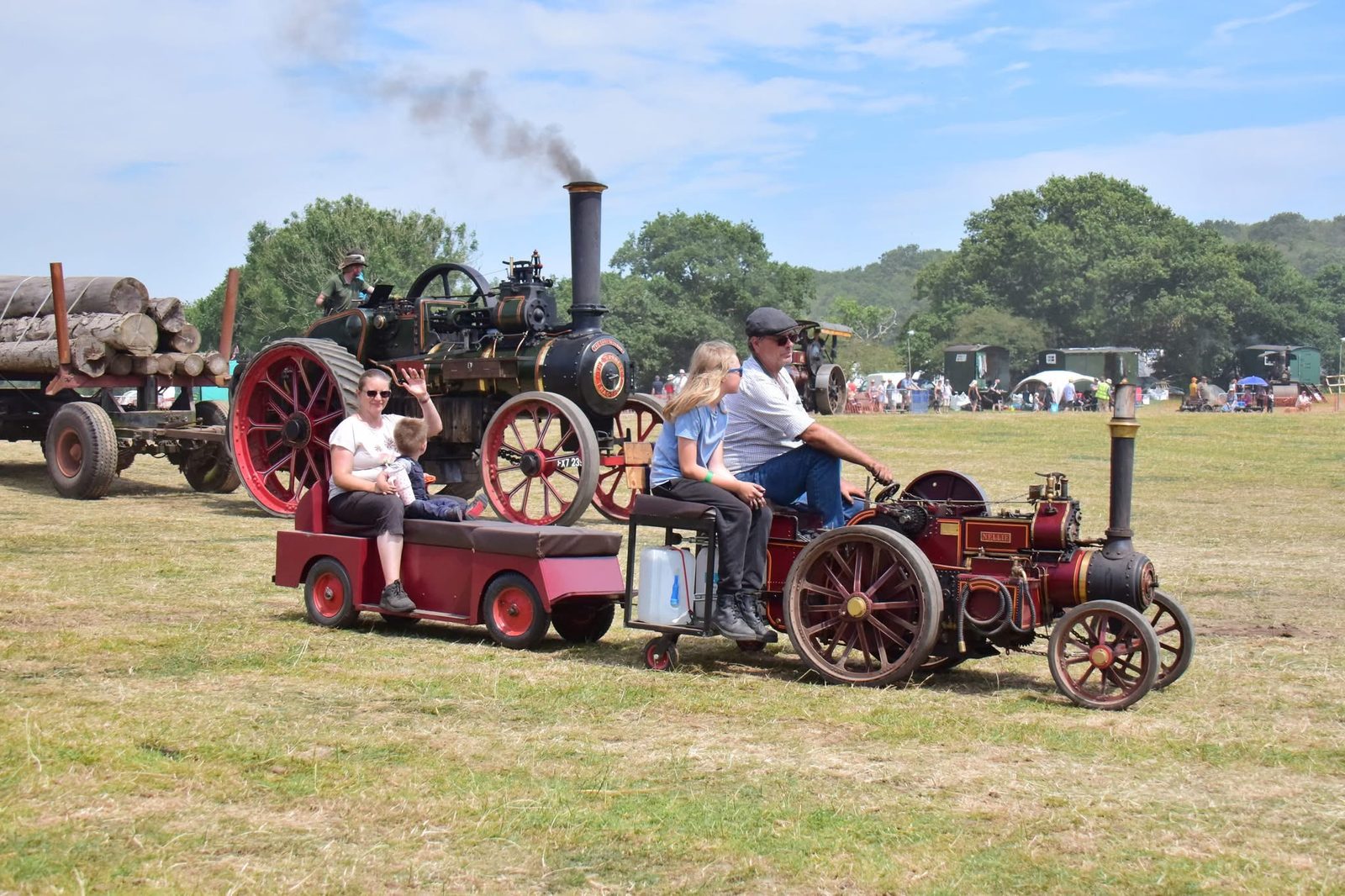Nellie hauling a trailer of passengers around the main ring at a steam rally, passing a full-size traction engine hauling timber